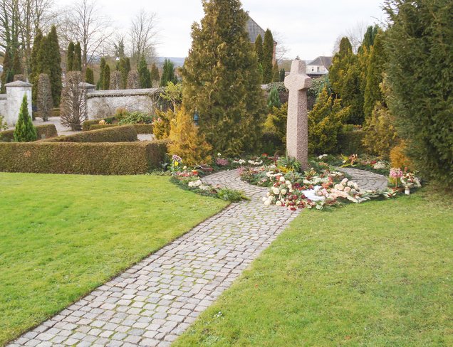 Gravplads med blomster foran stor kors-stenfigur.